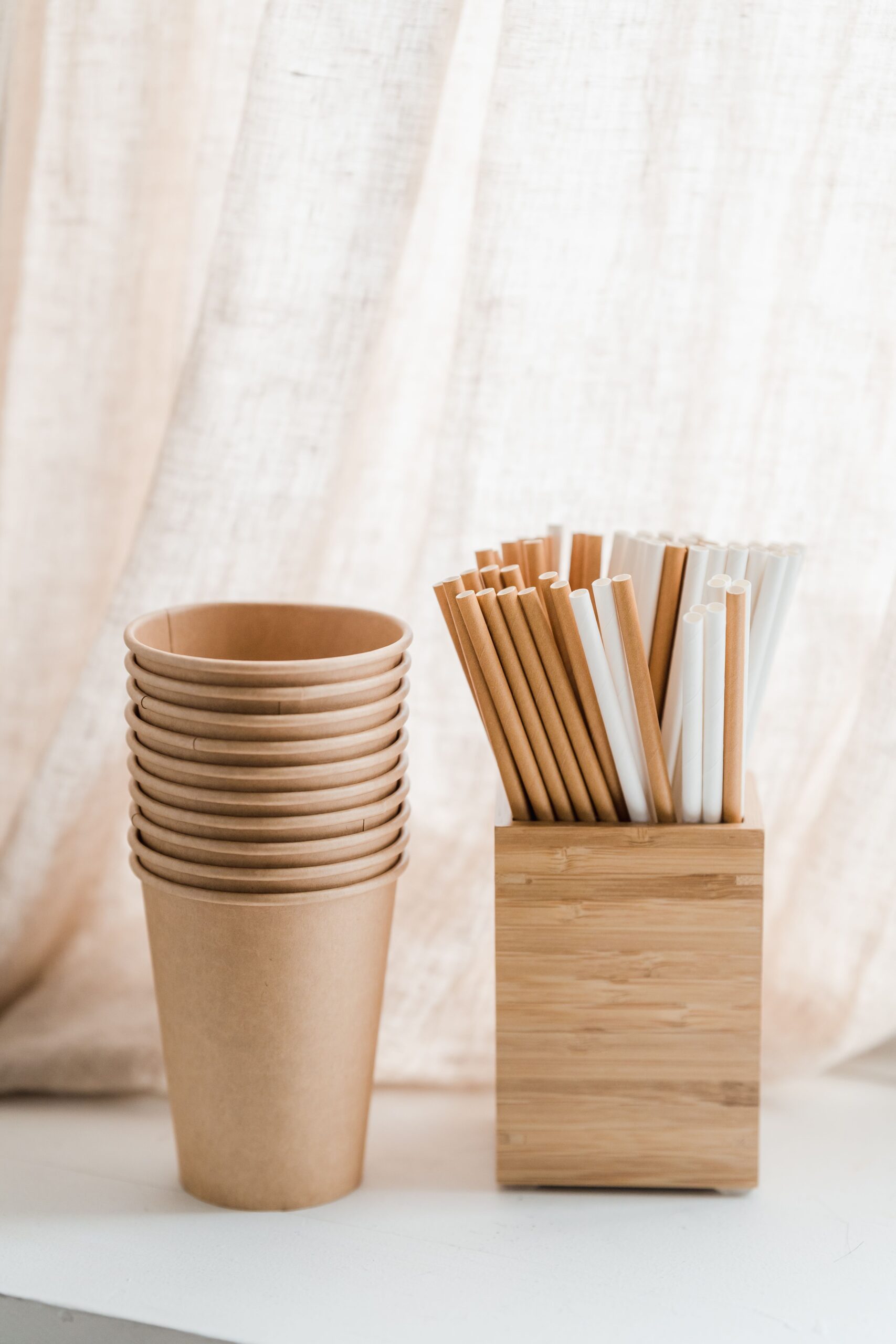 Stacked eco-friendly paper cups and bamboo straws organized in a wooden box against a soft fabric backdrop.